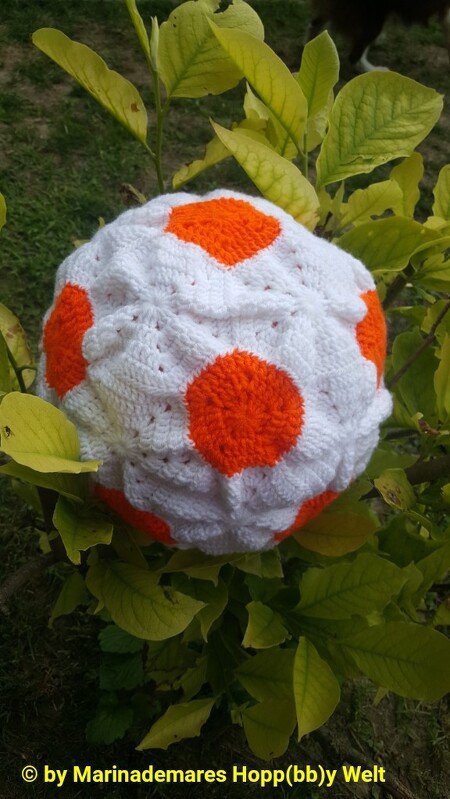 Rundes gehäkeltes Kissen in Weiß und Orange mit markantem Fußballmuster.