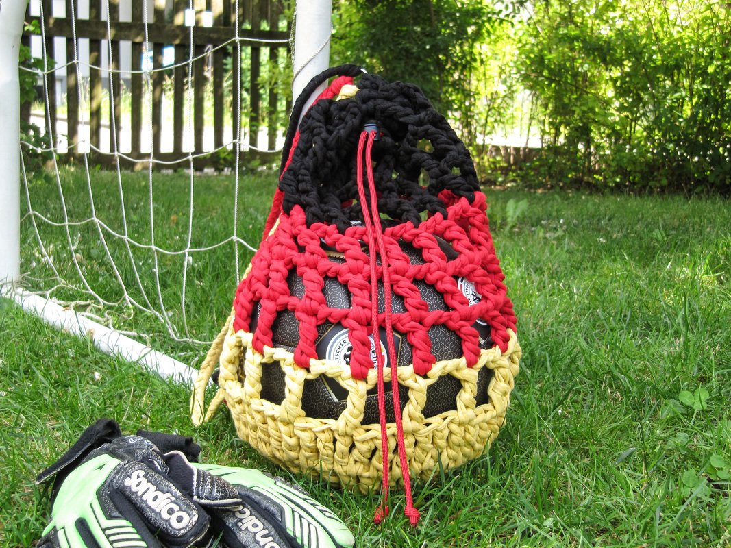 Handgehäkelte Netztasche in Schwarz‑Rot‑Gold mit Fußball, auf Rasen neben Fußballtor und Torwarthandschuhen.