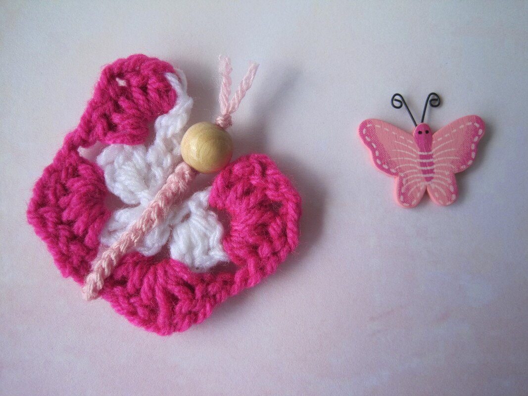 Gehäkelte Schmetterlingsapplikation in Pink und Weiß mit Holzperle und kleinem rosa Holzschmetterling auf hellem Untergrund.