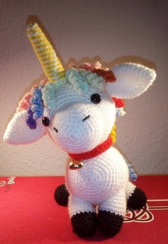 Weißes sitzendes Amigurumi-Einhorn mit schwarzen Hufen, gelb-weiß gestreiftem Horn, pastellfarbener Mähne und rotem Halsband mit Glöckchen