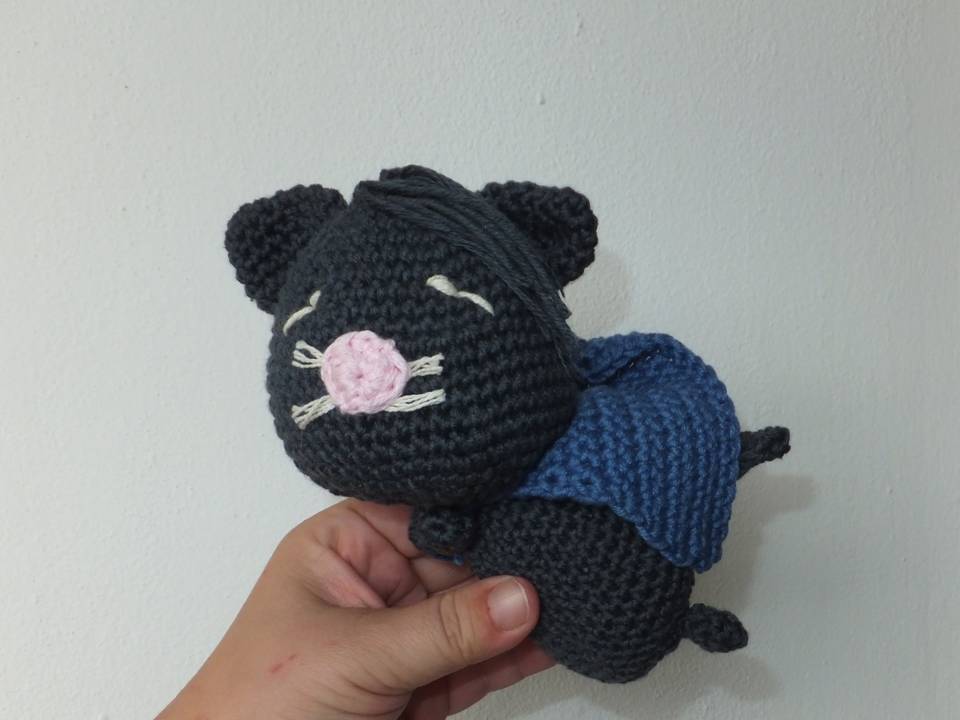 Hand hält dunkelgraue gehäkelte Amigurumi-Katze mit rosa Nase und blauem Umhang