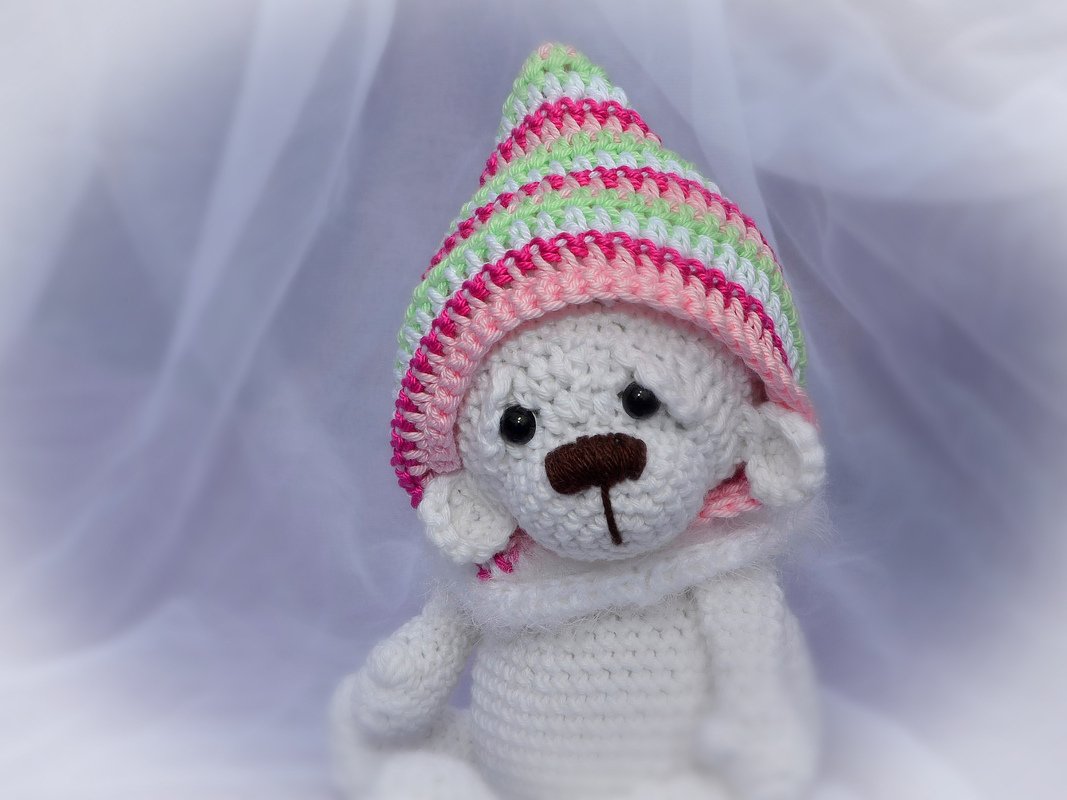 Weißer, handgehäkelter Teddybär mit pink-grün-weißer Zipfelmütze und weißem Schal vor hellem Hintergrund.