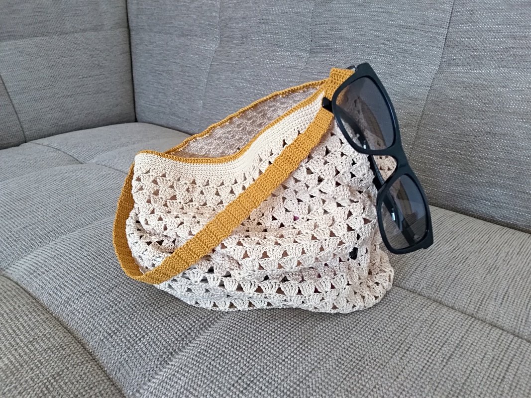 Offen gehäkelte beigefarbene Tasche mit feinem Lochmuster und senfgelbem Trageriemen auf grauem Sofa, schwarze Sonnenbrille daneben.