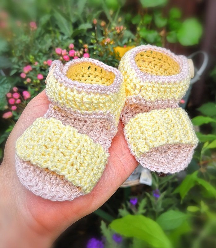 Hand hält gehäkelte Babysandalen in Pastellgelb und Rosa