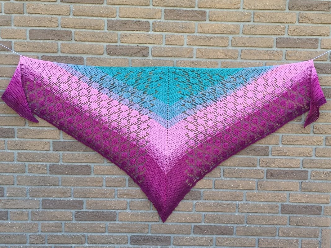 Gehäkeltes Dreieckstuch mit Farbverlauf in Magenta, Pink und Türkis mit durchbrochenem Muster, vor einer Ziegelwand gespannt.
