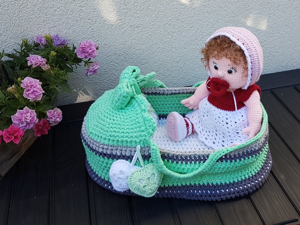 Gehäkelte Babypuppe mit rosa Haube im gestreiften Körbchen neben violetten Blumen.