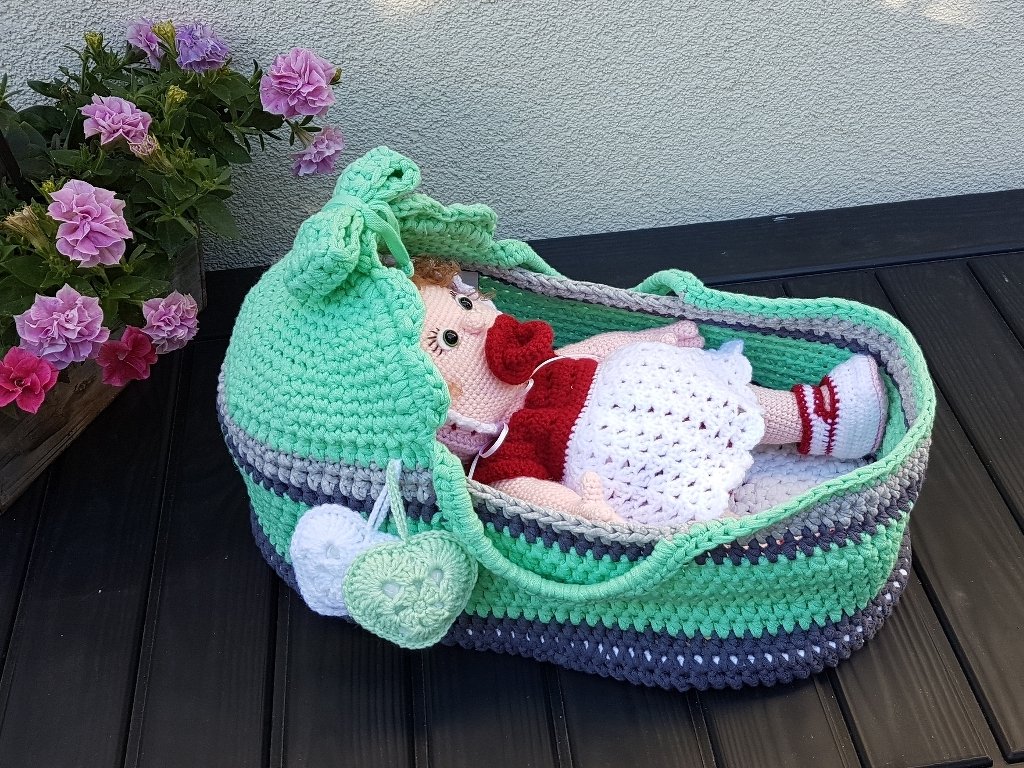Gehäkelte Puppe mit rotem Oberteil und weißem Kleid in mintgrün-grau gestreiftem Tragekorb, Blumen im Hintergrund