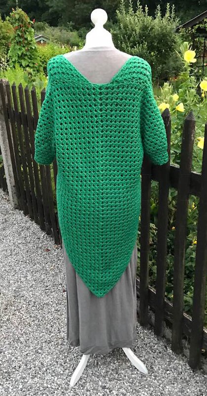 Rückansicht eines grasgrünen gehäkelten Poncho-Tops mit Lochmuster, tiefem V-Ausschnitt im Rücken und spitz zulaufendem Saum auf einer Schaufensterpuppe vor einem Holzzaun.