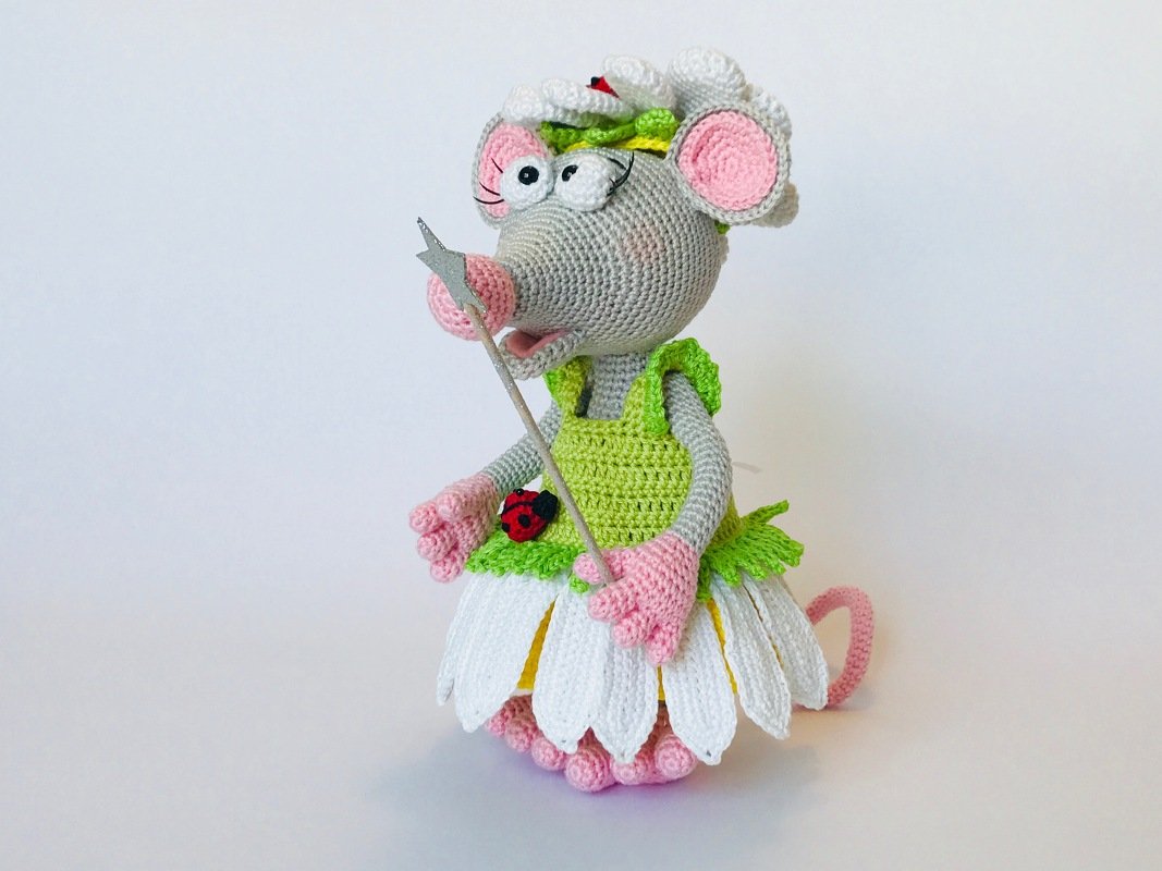 Gehäkelte graue Amigurumi-Maus mit pinken Ohren und Händen, grünem Oberteil, weißem Blütenrock, Marienkäfer und silbernem Zauberstab.