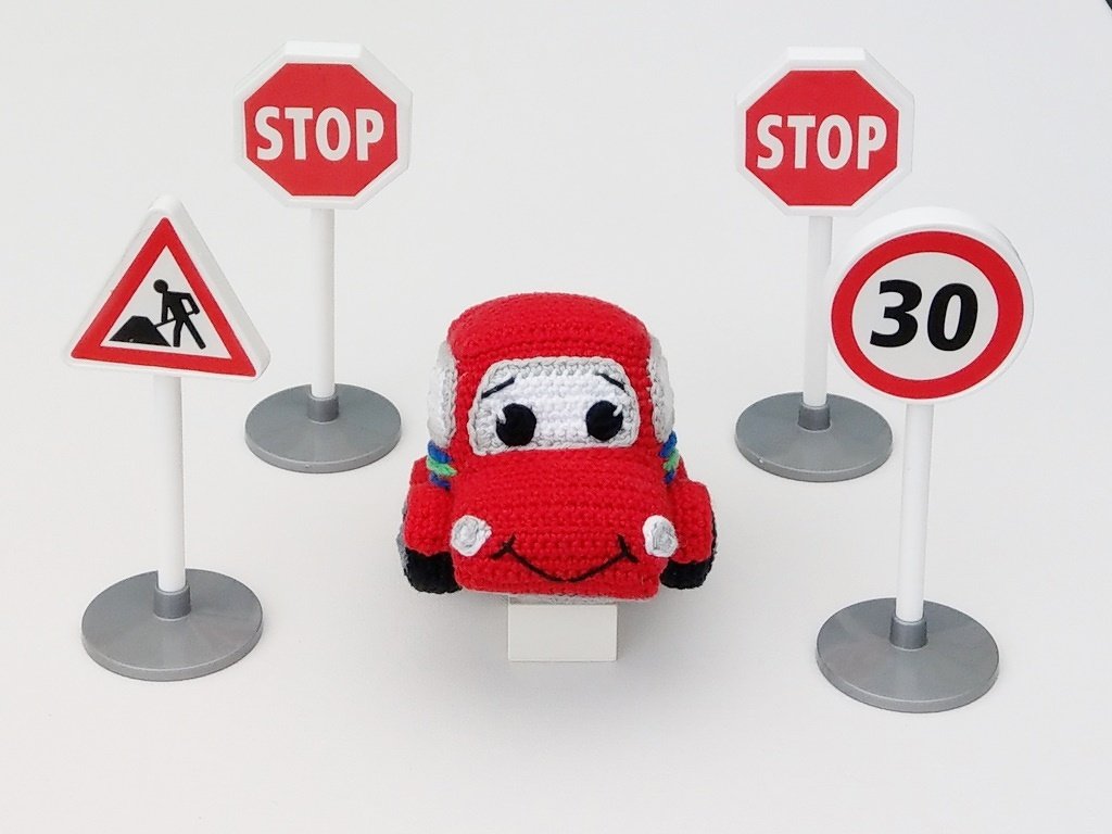 Rotes gehäkeltes Amigurumi-Auto umgeben von vier Spielzeug-Verkehrsschildern auf weißem Hintergrund.