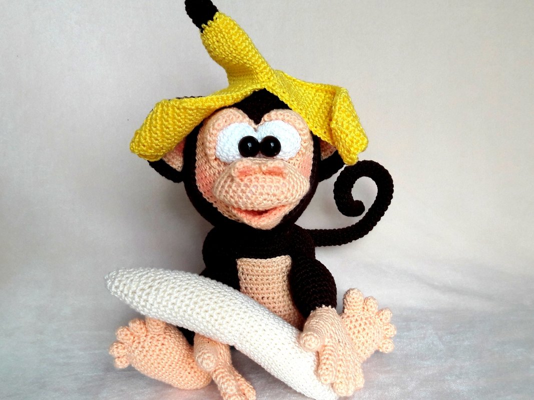 Crocheted amigurumi monkey, dark brown with peach face and a yellow banana peel hat holding a white crocheted banana.