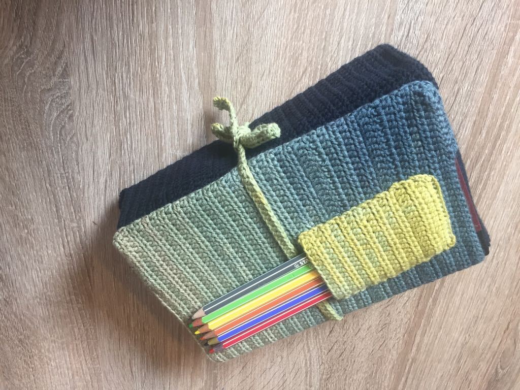 Drei gehäkelte Stiftetuis in Blau- und Grüntönen, zusammengebunden, eines mit farbigen Buntstiften, auf Holzuntergrund.