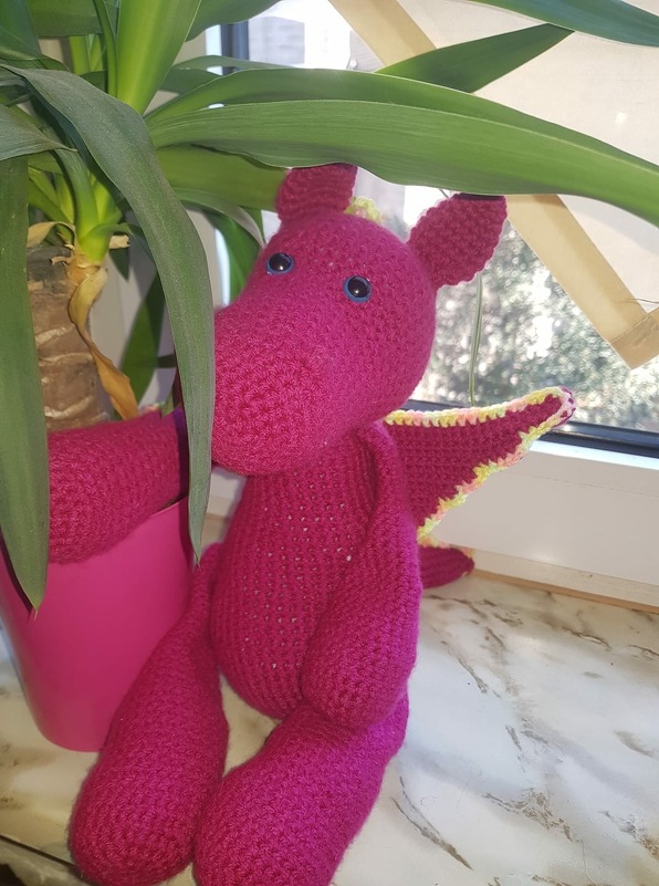 Pinker gehäkelter Drache mit blauen Augen und hell gerandeten Flügeln neben einer Zimmerpflanze auf einer Fensterbank