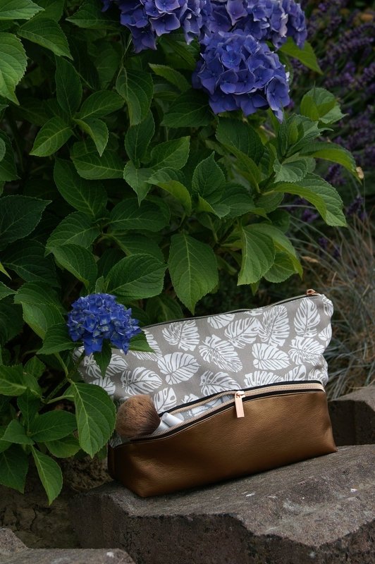 Beige Kosmetiktasche mit weißem Blattmuster und braunem Lederoptik-Boden auf Stein vor blauen Hortensien
