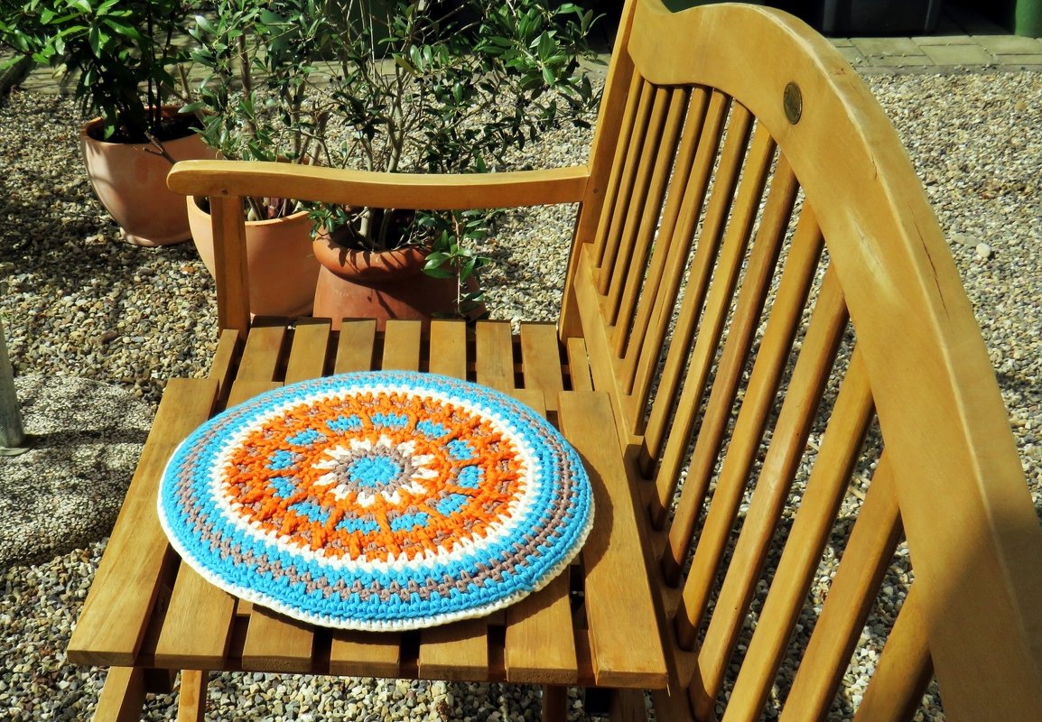 Rundes gehäkeltes Kissen in Orange, Türkis, Weiß und Grau mit Mandala-Muster auf einem Holzstuhl