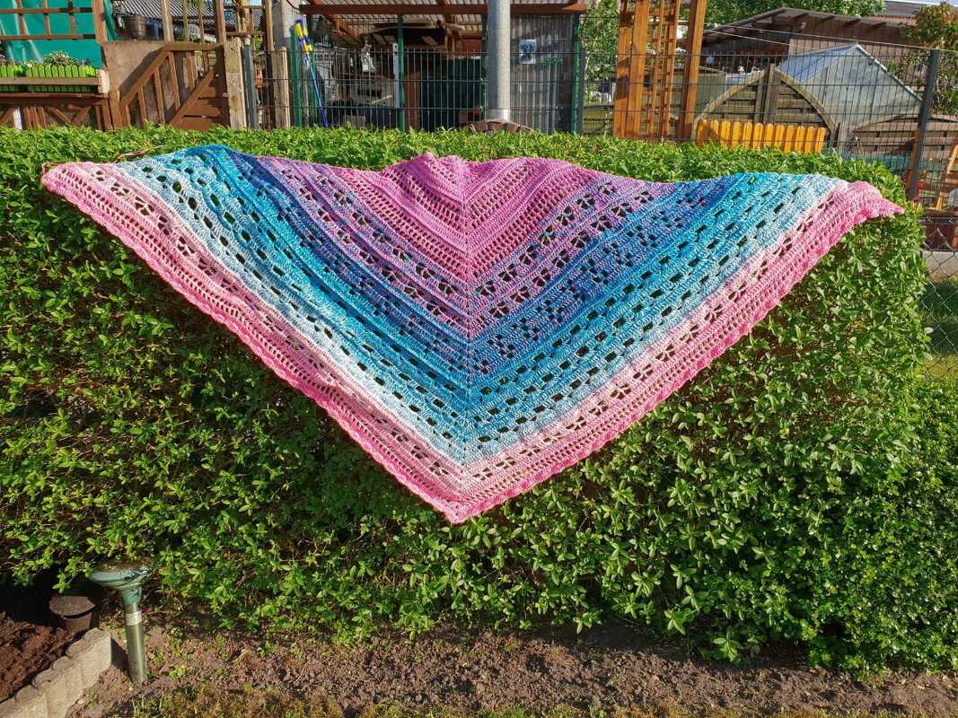 Gehäkeltes Dreieckstuch mit Farbverlauf in Rosa, Violett und Blau, drapiert über einer grünen Hecke