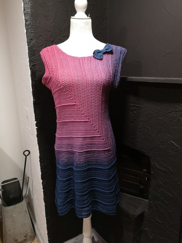 Ärmelloses gehäkeltes Damenkleid mit Pink‑Blau‑Farbverlauf und kleiner blauer Schleife an der Schulter auf Schneiderpuppe.