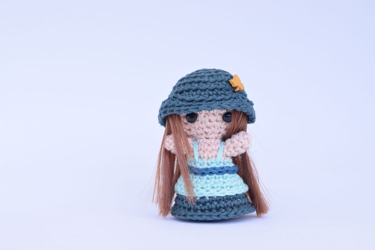Gehäkelte Amigurumi-Puppe mit braunen Haaren, zweifarbigem blauem Kleid und dunkelblauem Hut mit gelbem Detail.