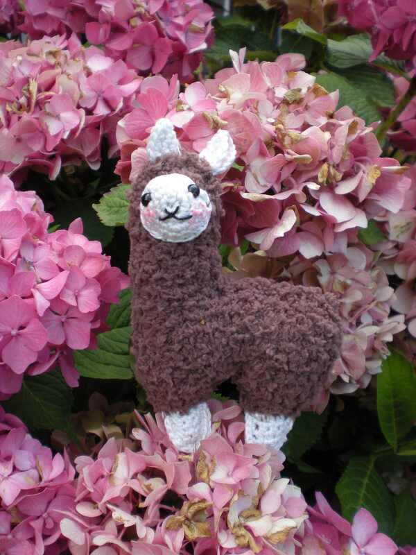 Brown crochet amigurumi llama with white face, pink blush, and white feet among pink hydrangea flowers.