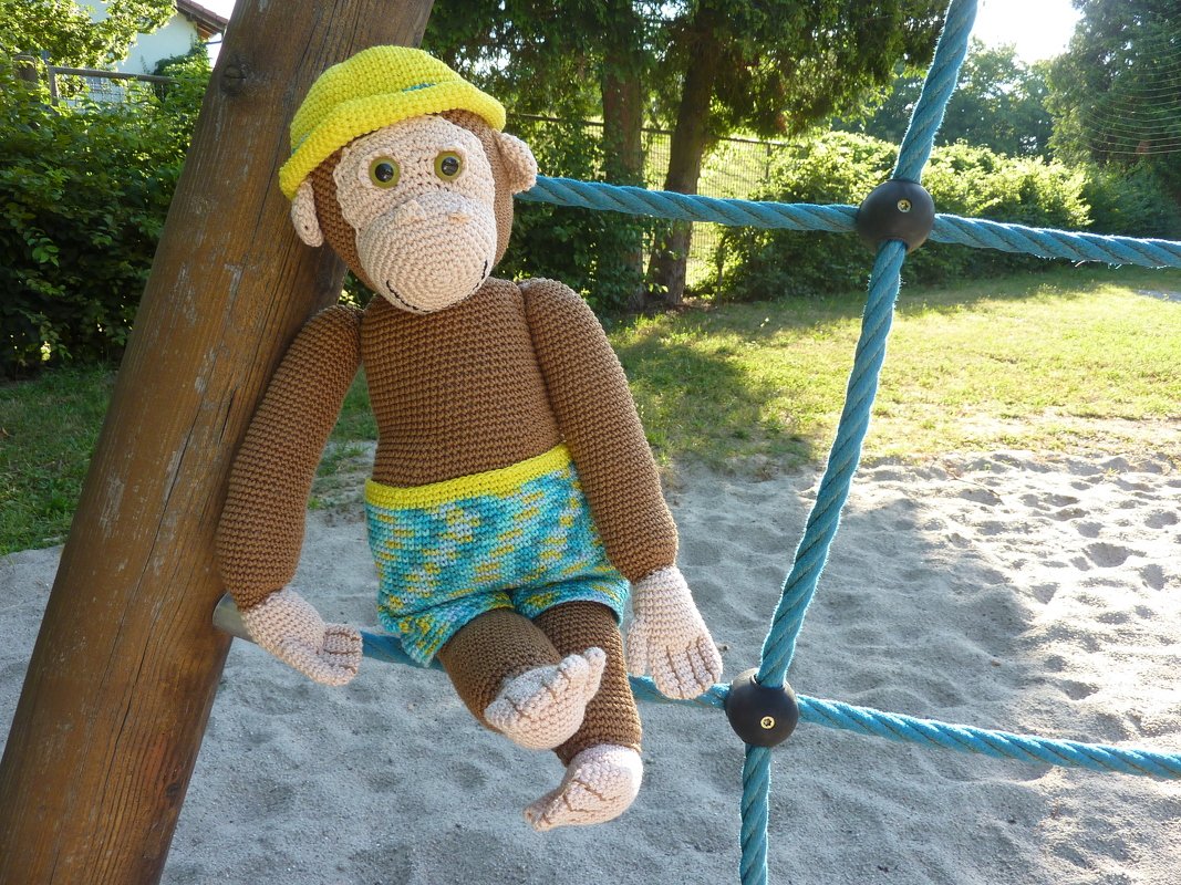 Gehäkelter Amigurumi-Affe mit gelbem Hut und bunten Shorts auf Kletternetz