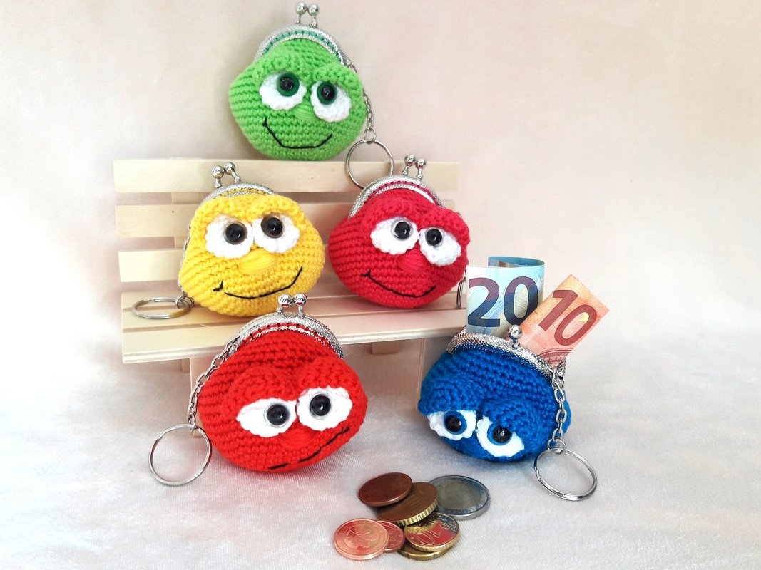 Five smiling crochet coin purses in green, yellow, red, and blue with metal kiss clasps and keyrings, arranged on a small stand with coins nearby.