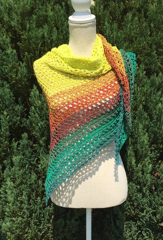 Beigefarbene Schneiderbüste zeigt ein gehäkeltes Dreieckstuch mit gelb‑orange‑grünem Farbverlauf vor Pflanzen