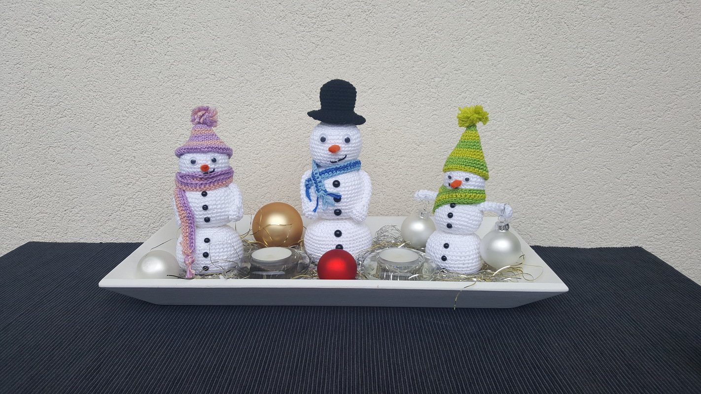 Drei gehäkelte Amigurumi-Schneemänner mit Mützen und Schals auf einem dekorierten Tablett.