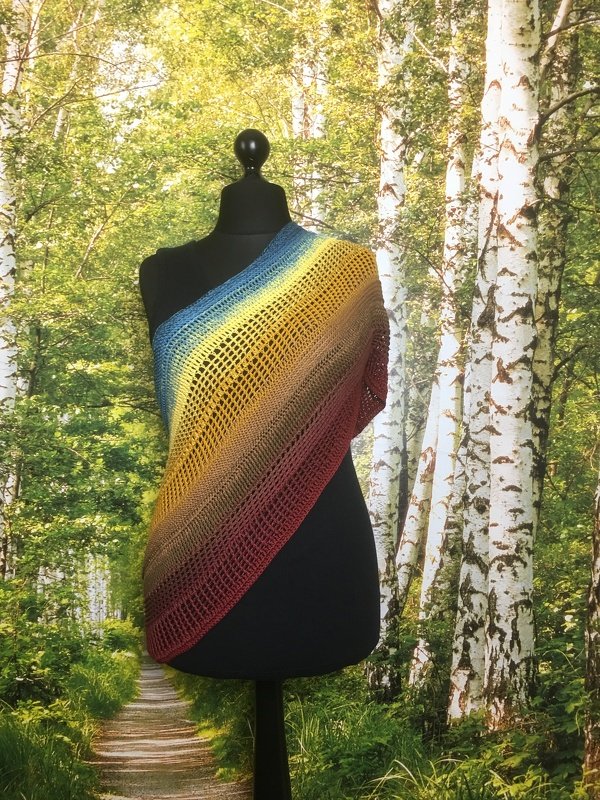 Gehäkeltes Schultertuch mit Farbverlauf (Blau, Gelb, Rotbraun) auf schwarzer Schneiderpuppe vor Birkenwald