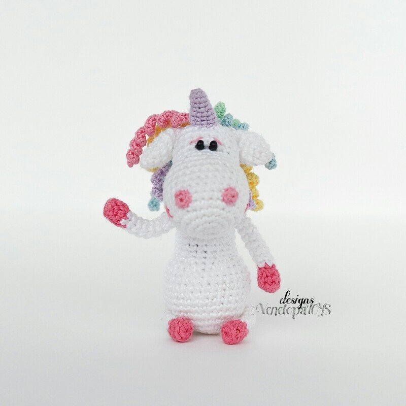 White crochet amigurumi unicorn with pastel curly mane, lilac horn and pink snout and hooves on white background