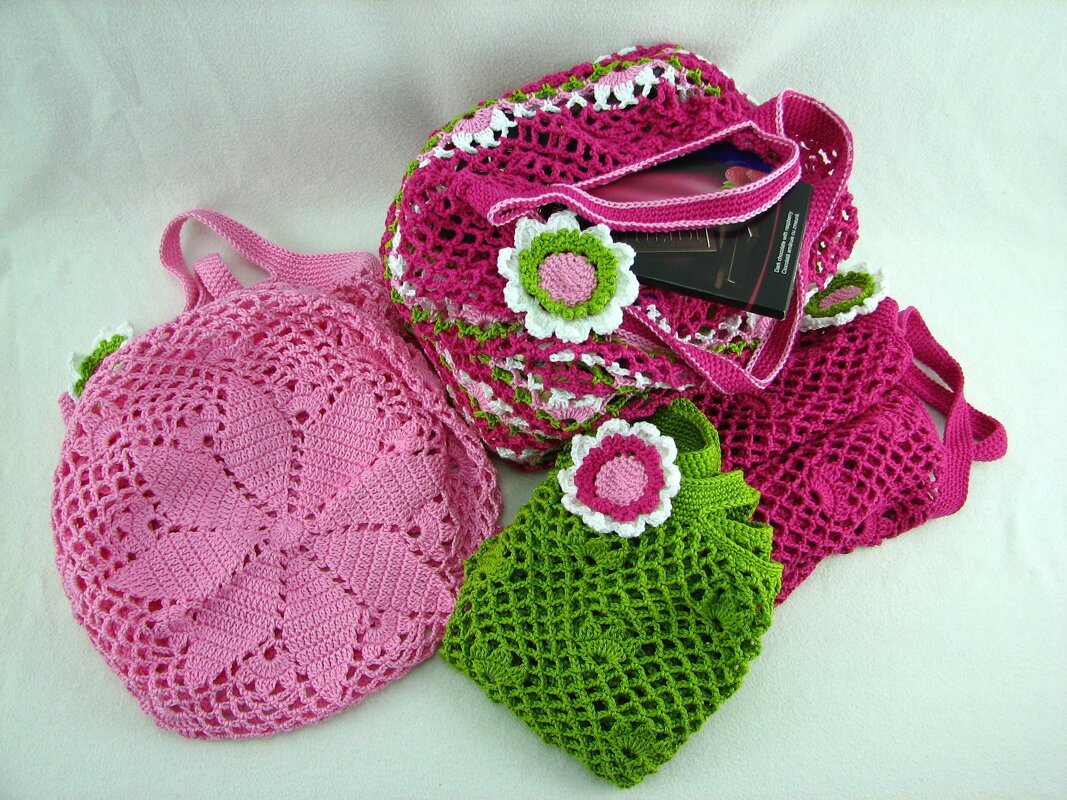 Drei gehäkelte Taschen: große fuchsiafarbene Henkeltasche, runde hellrosa Tasche und kleine grüne Beuteltasche mit Blumenapplikationen.