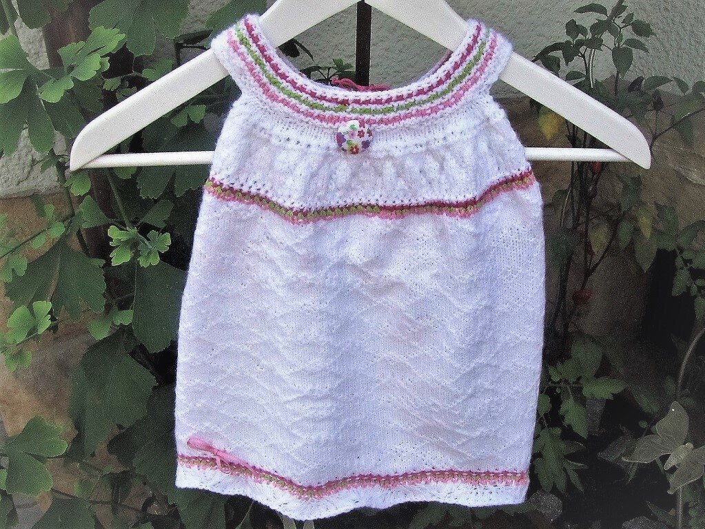 Weißes gestricktes Babytop mit rosa und grünen Streifen und Zickzackmuster, an einem Bügel vor grünen Pflanzen hängend.