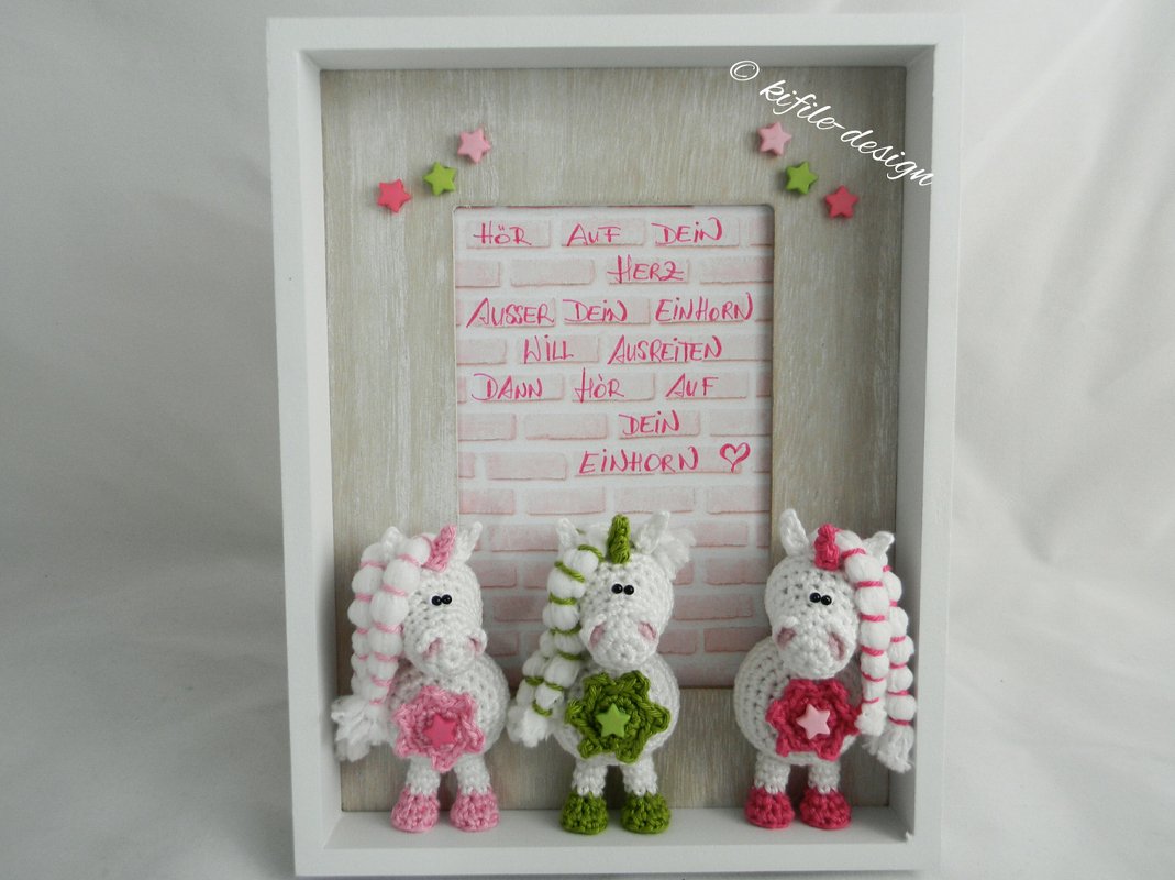Weißer Rahmen mit drei gehäkelten Amigurumi‑Einhörnern vor rosafarbenem Ziegel‑Hintergrund und handgeschriebenem Spruch.