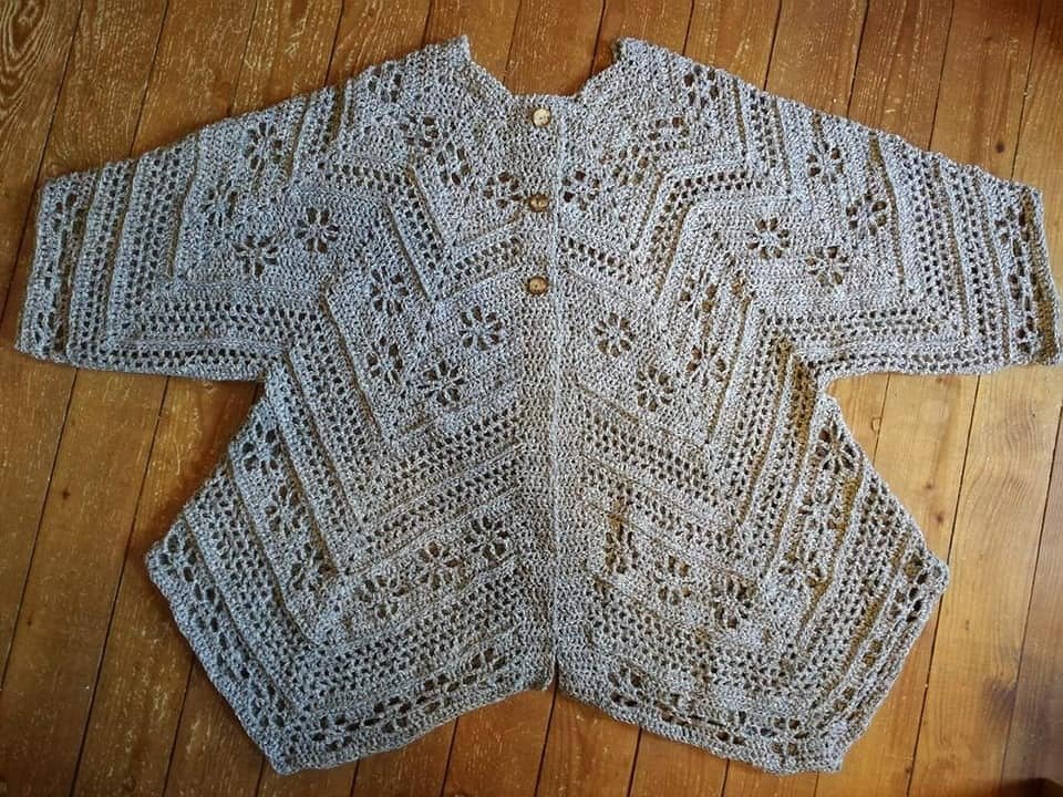 Gehäkelte Damenjacke in Grau-Beige mit Zacken- und Blütenmuster und drei Holzknöpfen, liegend auf Holzfußboden.