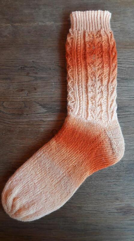 Einzelne handgestrickte Socke mit Orange-Creme-Farbverlauf, Zopf- und Blattmuster am Schaft, geripptes Bündchen auf Holzboden