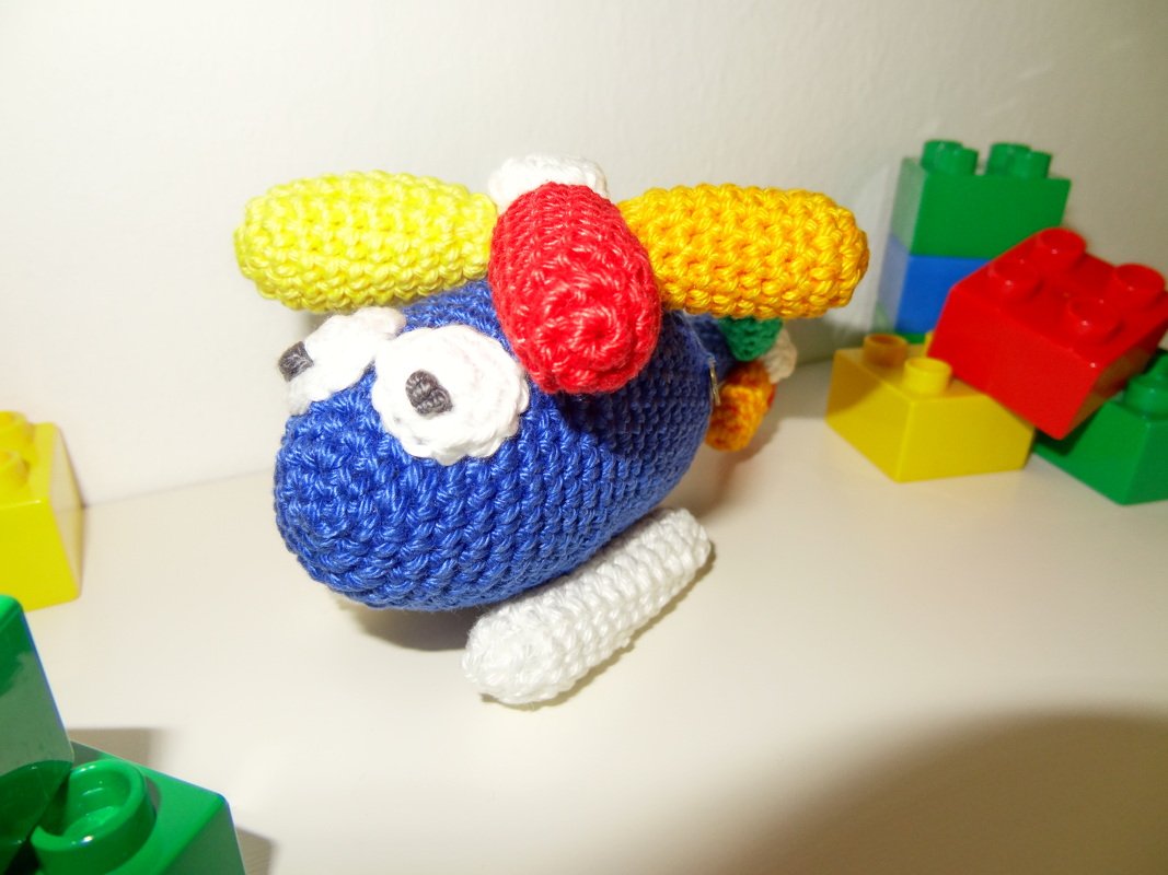 Blauer gehäkelter Amigurumi-Hubschrauber mit weißen Augen und gelben sowie roten Rotoren, umgeben von bunten Bausteinen.