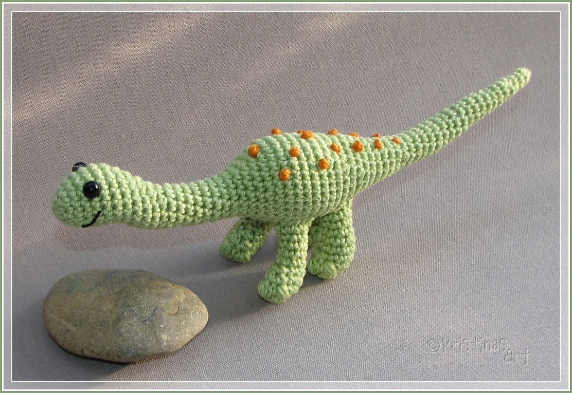 Hellgrüner gehäkelter Amigurumi-Dinosaurier mit orangen Noppen, schwarzen Augen und gesticktem Lächeln neben einem flachen Stein