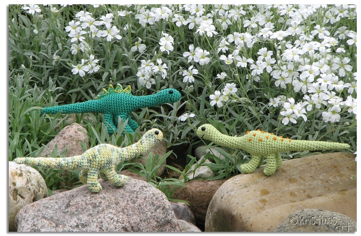 Drei gehäkelte Dinosaurier auf Felsen vor weißen Blüten