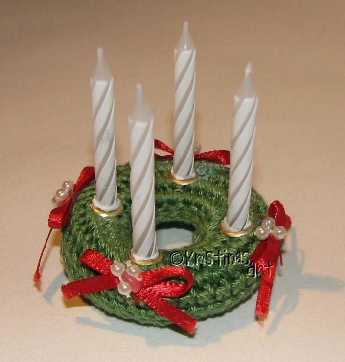 Gehäkelter grüner Mini-Adventskranz mit vier weißen Kerzen, roten Schleifen und Perlen.