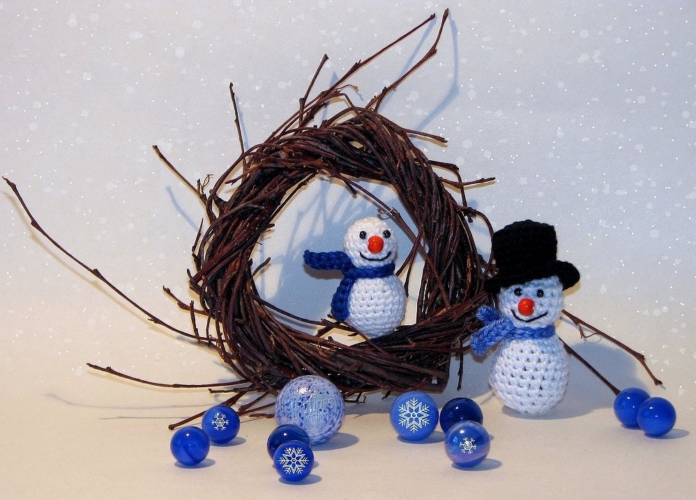 Zwei gehäkelte Schneemänner, einer mit Zylinder, vor einem Zweigkranz und blauen Kugeln