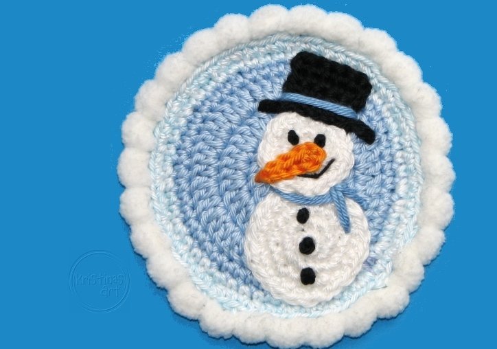 Runde gehäkelte Schneemann-Applikation mit schwarzem Zylinder, orangefarbener Nase und blauem Schal auf hellblauem Hintergrund mit weißem Bommelrand.