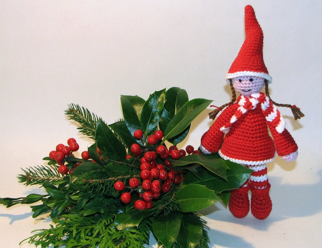 Rote gehäkelte Amigurumi-Puppe mit Zipfelmütze neben Stechpalmenzweig und roten Beeren