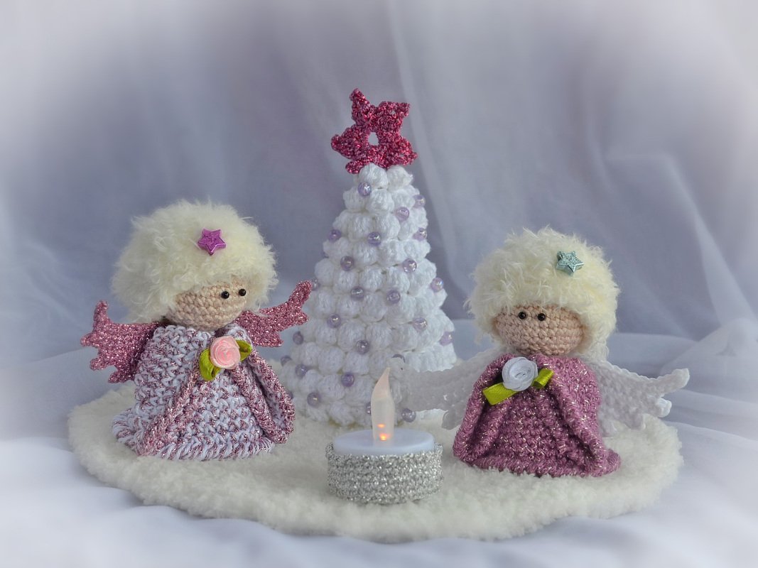 Zwei gehäkelte Amigurumi-Engel mit glitzernden Flügeln und ein weißer Häkel-Weihnachtsbaum mit Stern auf flauschiger Unterlage, beleuchtet von einem LED-Teelicht.