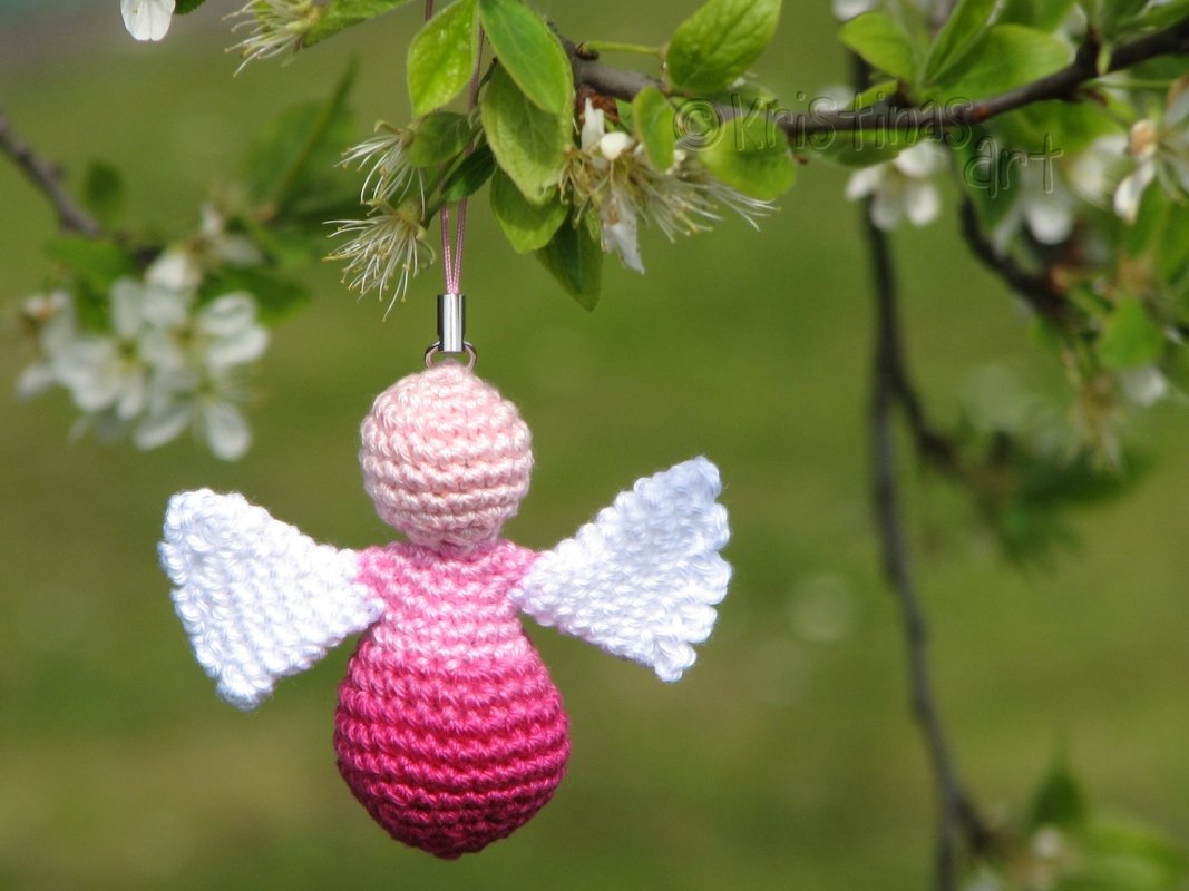 Gehäkelter Engel in Pink mit weißen Flügeln an blühendem Zweig hängend.