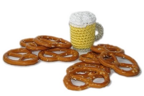 Kleiner gehäkelter gelber Bierkrug mit weißer Schaumkrone neben mehreren kleinen Brezeln mit Salz.