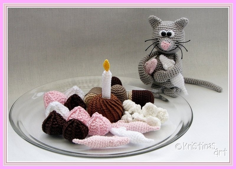 Gehäkelte graue Katze neben einer Glasplatte mit Kuchen, Kerze und kleinen Häkelbonbons in Rosa, Braun und Weiß.