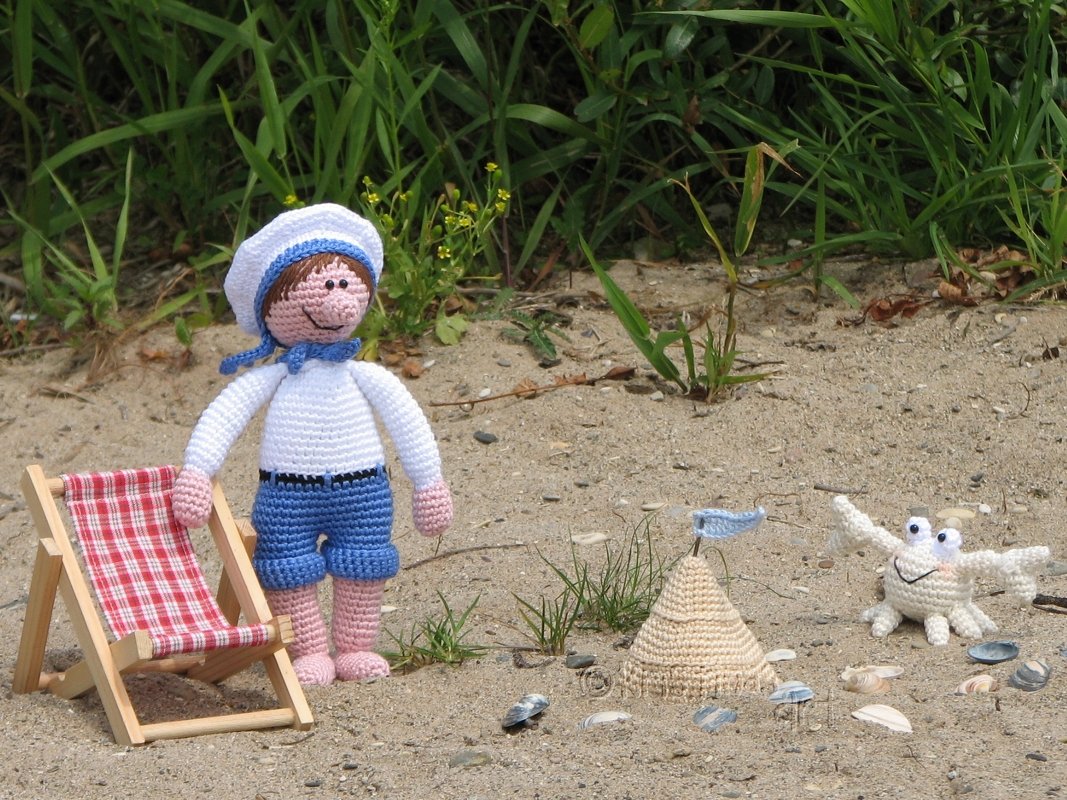 Gehäkelte Matrosenpuppe neben Liegestuhl, Sandburg und Amigurumi-Krabbe auf Sand