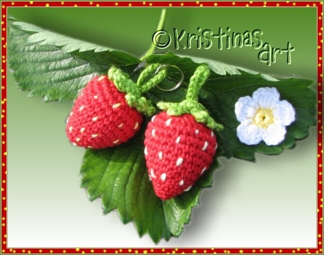 Nahaufnahme zweier gehäkelter Erdbeeren mit weißen Stichen, grünen Kappen, Metallringen und einer kleinen weißen Häkelblüte