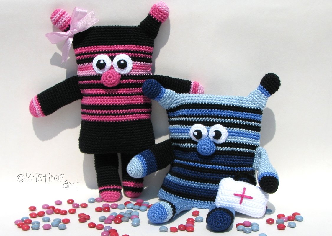 Zwei gestreifte Amigurumi-Figuren in Rosa‑Schwarz und Blau‑Schwarz, eine mit Schleife, eine mit kleinem Täschchen mit rotem Kreuz, auf hellem Hintergrund mit Süßigkeiten.