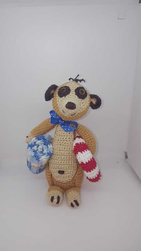 Gehäkeltes Amigurumi-Erdmännchen, braun-beige, mit blauer Schleife, blau-weißer Tasche und rot-weißem Objekt