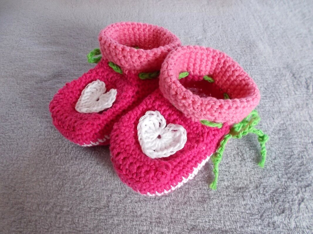 Zwei gehäkelte Babyschuhe in Pink mit weißen Herzapplikationen und hellrosa Bündchen auf grauem Untergrund.